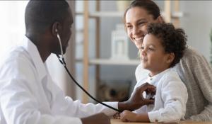 A young black doctor holds a stethoscope up to a mixed child's heart. The mom has the child on her lap. Both are smiling at the doctor.